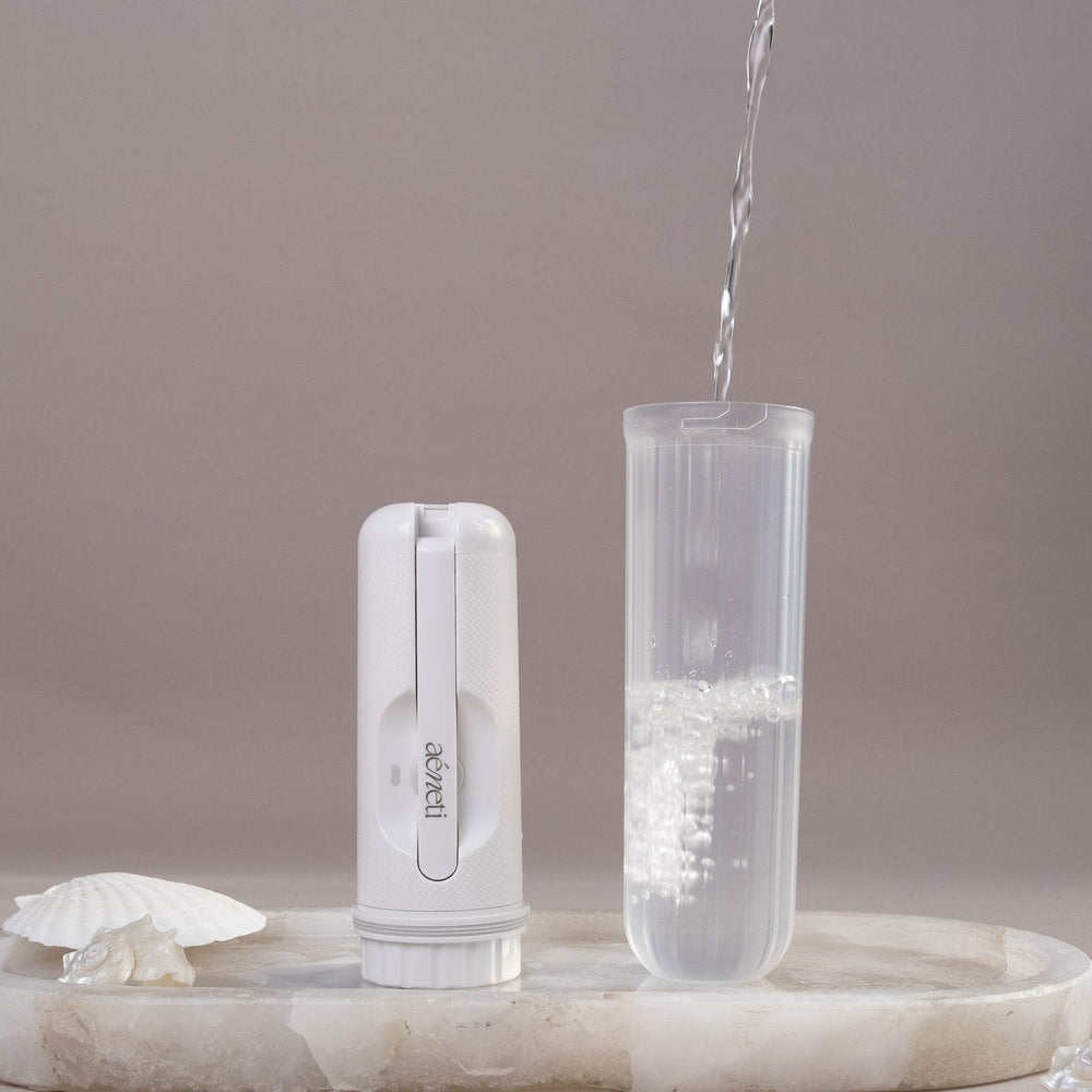 A white handheld portable bidet stands next to a clear cylindrical container being filled with water, set on a light marble tray with seashell props against a soft taupe background.