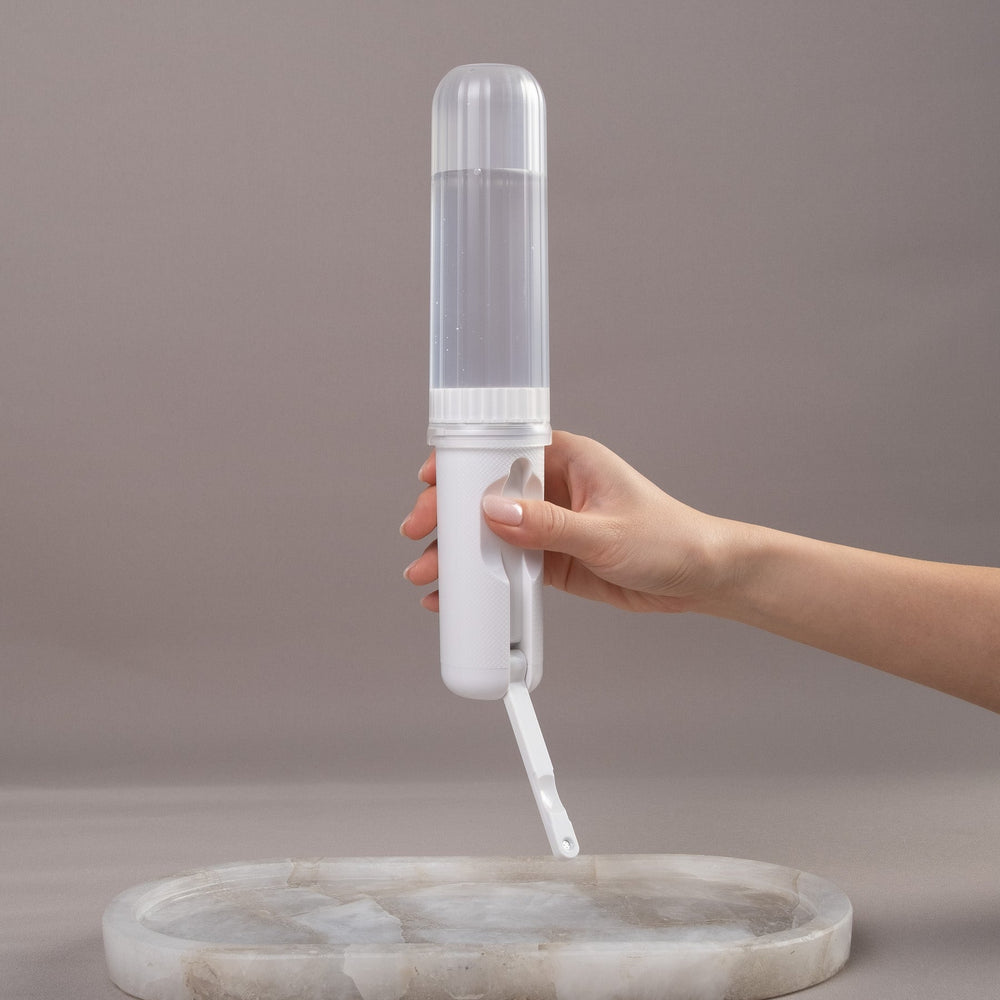 A hand holding a white and clear portable bidet with a flip-up nozzle, positioned above a round white marble tray on a neutral gray background.