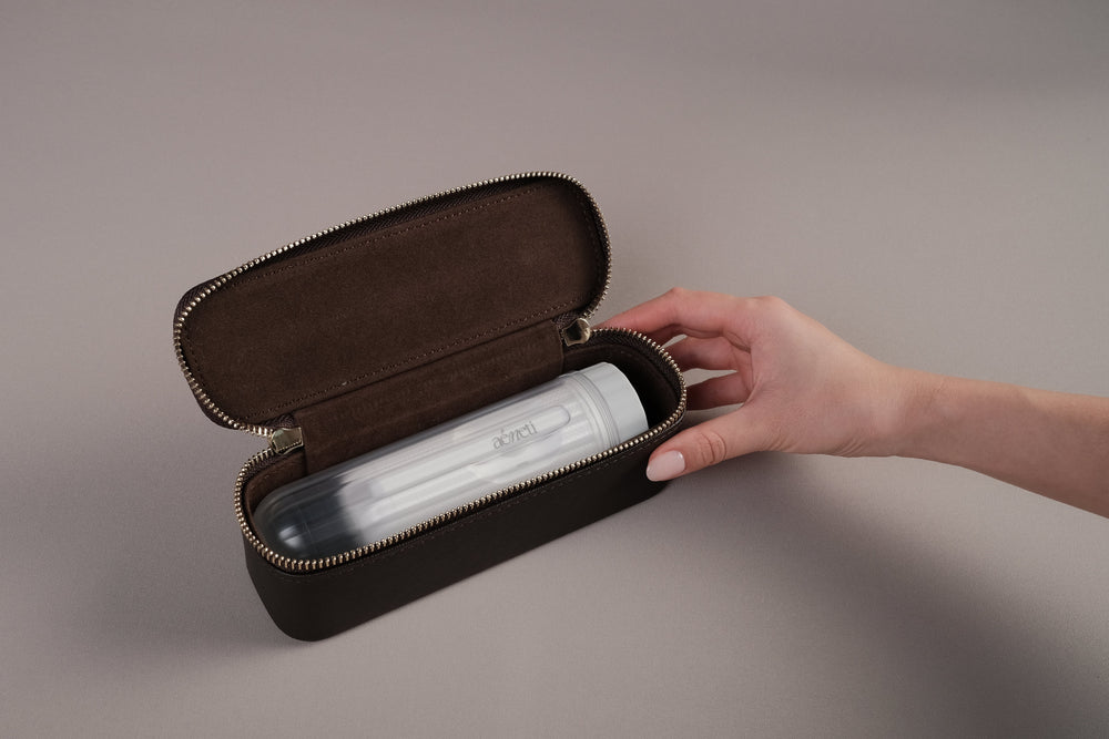 A hand is shown placing a white portable bidet into an open dark brown zippered case with a brown interior, set against a neutral gray background.