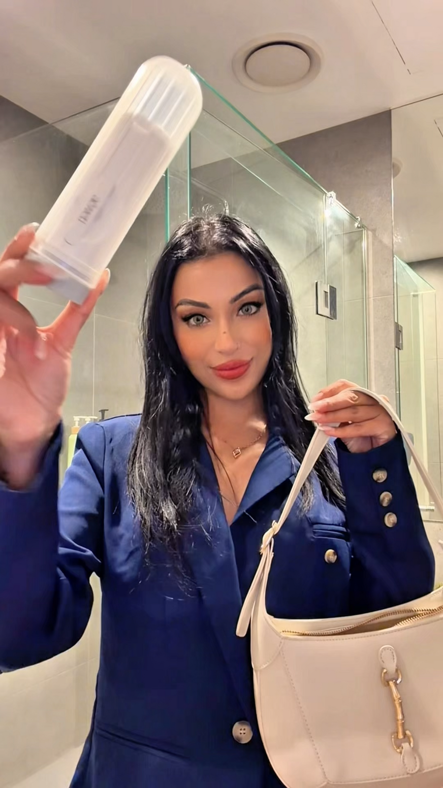A woman in a navy blue blazer holds up aeneti portable bidet in her right hand and carries a cream-colored shoulder bag in her left hand, photographed in a bathroom setting.